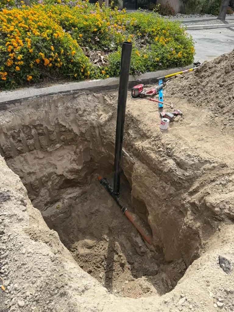 Deep square excavation beside a flowering hillside with a pipe standing in the pit