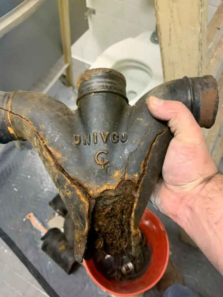 Plumber holding a cracked cast-iron UNIVCO closet wye packed with scale, bathroom rebuild behind