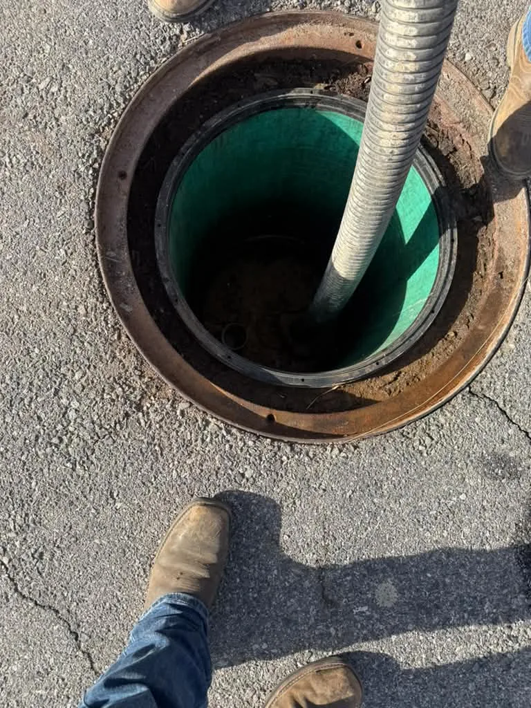 Suction hose lowered into a green-lined vault through a manhole, crew boots at the rim