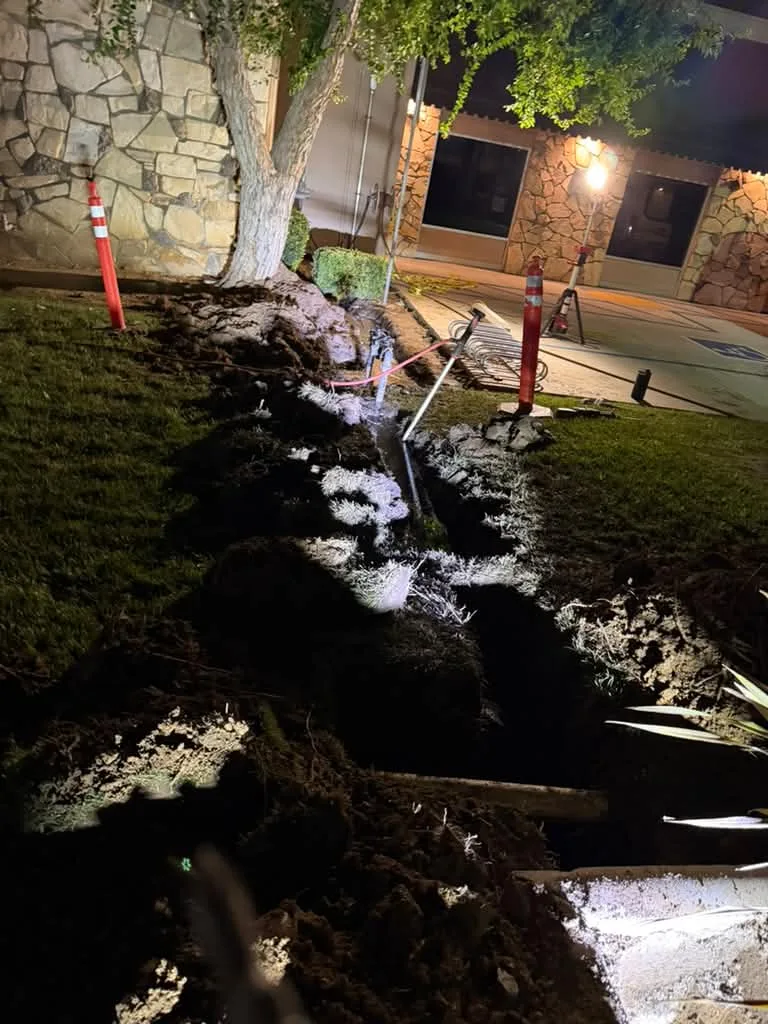 Night excavation at a street curb under work lights, water pooling in the cut
