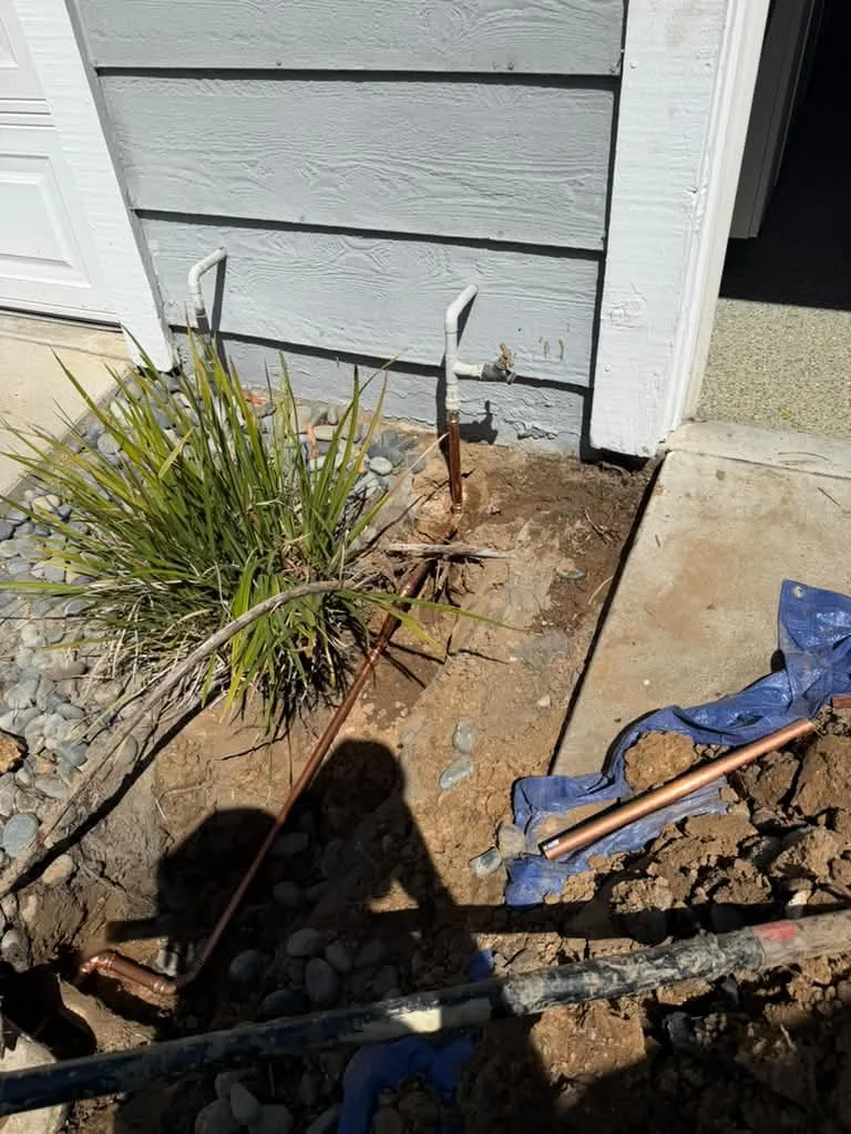 Open excavation along a stucco house side yard, saturated dark soil, pipe and tools laid out